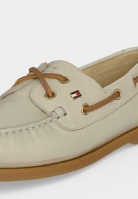Μπεζ δερμάτινο boat shoe με δερμάτινους κορδόνια σε ταμπ χρώμα που περνούν από χρυσά μεταλλικά ανοίγματα και μικρό ορθογώνιο λογότυπο στο πλάι.