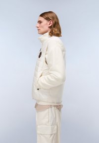 Hellfarbige, Reißverschlussjacke mit gesteppter Vorderseite, hohem Kragen und Seitentaschen. Mit einem runden Logo-Detail auf der Brust.