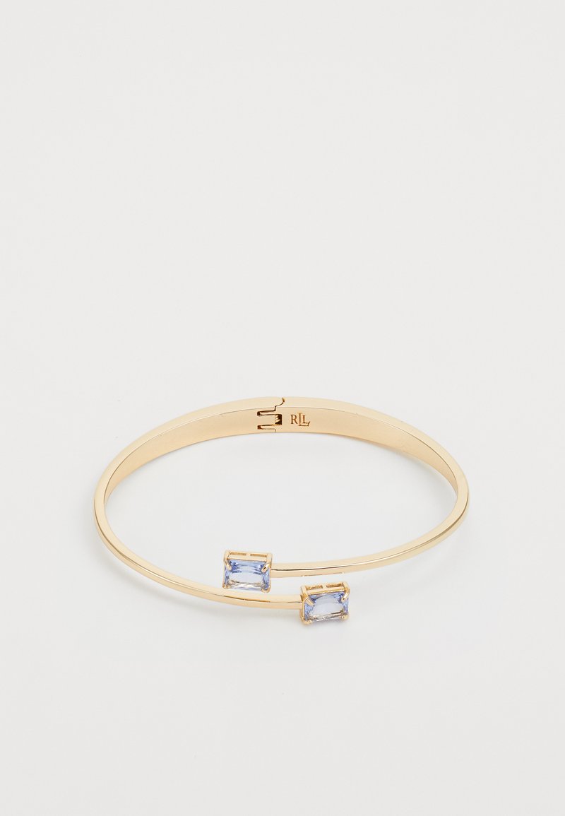 Zlatni metalni bangle z dvema pravokotnima svetlo modrima draguljema. Gladka površina z vgraviranim logotipom na notranji strani.