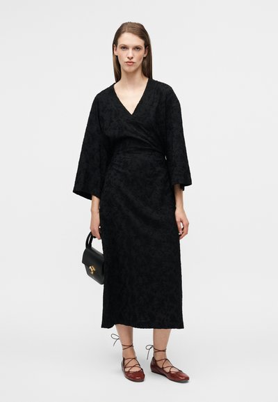 Robe midi noire à motifs floraux avec un col en V, des manches larges et une taille cintrée, accompagnée d'un sac à main noir et de ballerines marron à lacets.