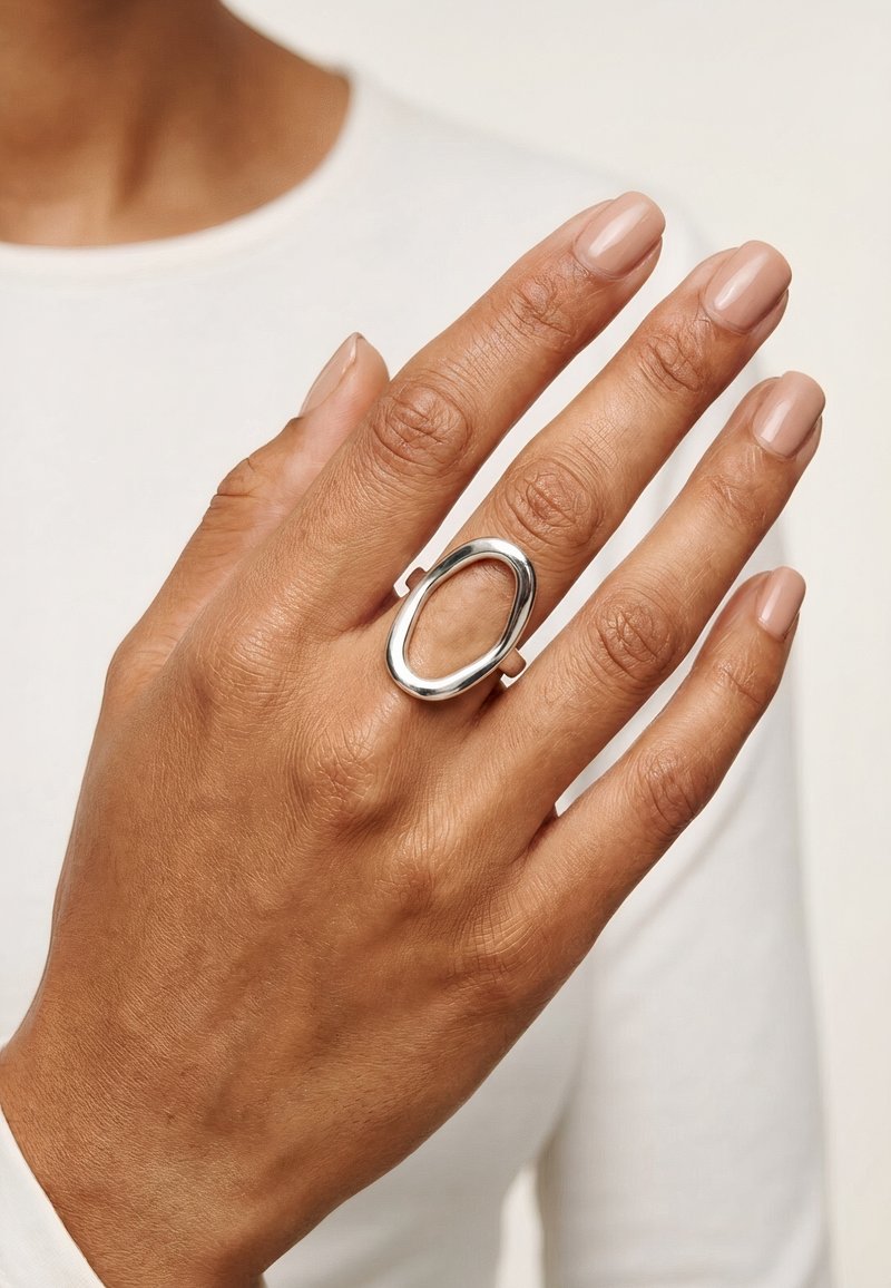 Main avec des ongles aux tons naturels portant une bague ovale ouverte en argent poli au majeur, sur un haut blanc à manches longues.