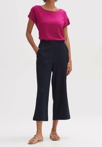 Fuchsiová blůza s kulatým výstřihem, v kombinaci s námořnickými culottes kalhotami. Modelka nosí ploché sandály; outfit ukazuje uvolněný, moderní siluetu.