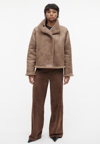 Bruine schapenvachtjas met een hoge kraag, gestructureerde stof en een knoopsluiting, gecombineerd met bruine wijde broek en zwarte puntige schoenen.