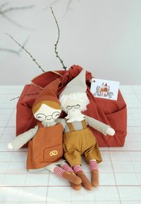 Dos muñecas de tela con características distintivas están sentadas junto a un regalo envuelto en tela de color óxido. Una lleva un sombrero y delantal rojos, la otra viste ropa gris y marrón.