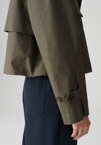 Olijfgroene cropped jack met een gelaagde achterkant, voorzien van een knoopmanchet en textiel met structuur, gecombineerd met marineblauwe broek.