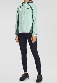 Veste de sport légère teal avec capuche et accents gris, dotée d'une fermeture éclair à l'avant et d'un logo. Associée à des leggings noirs et des chaussures de course.