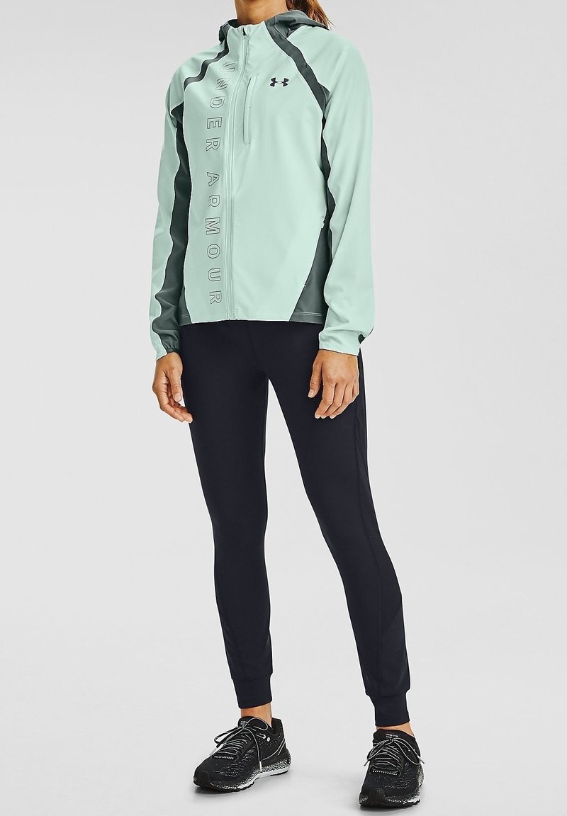 Veste de sport légère teal avec capuche et accents gris, dotée d'une fermeture éclair à l'avant et d'un logo. Associée à des leggings noirs et des chaussures de course.