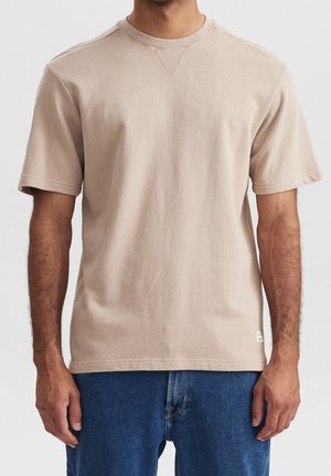 Homme portant un t-shirt beige clair à manches courtes et col rond, ainsi qu'un jean bleu, visible du cou jusqu'aux cuisses sur un fond uni.