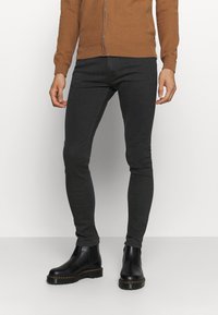 Man i svarta slim-fit byxor, svarta läder Chelsea-boots och en brun zip-up tröja, stående mot en enkel vit bakgrund.