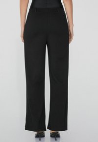 Pantalones de pierna ancha negros con una textura de tela suave, cintura alta y sutiles bolsillos laterales. Presenta costuras a medida y un ajuste relajado.