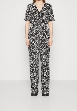 Vrouw draagt een zwart-witte jumpsuit met abstract patroon, korte mouwen, V-hals, getailleerde taille en zwarte schoenen met dikke zolen.