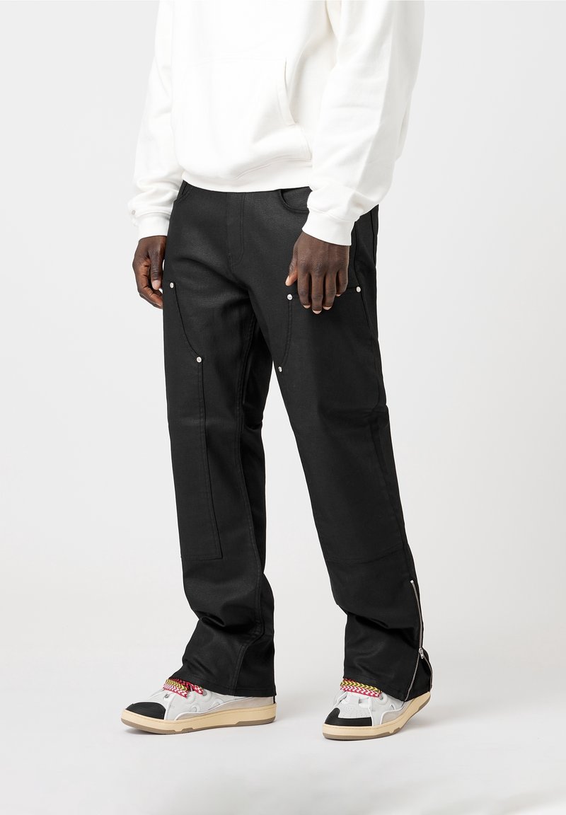 Homme portant un pantalon cargo noir avec des fermetures éclair aux chevilles et des baskets blanches à lacets multicolores, debout devant un fond uni.