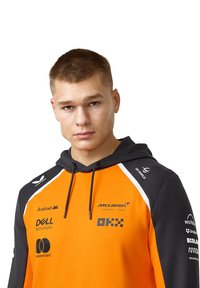 Pomarańczowa i czarna bluza z kapturem z logo McLaren, z białymi akcentami i logo różnych sponsorów nadrukowanymi z przodu.