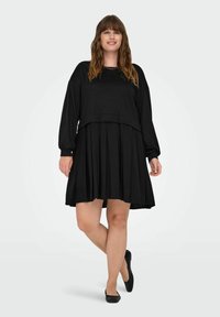 Abito nero con gonna a pieghe e top oversize a maniche lunghe. Tessuto morbido, vestibilità rilassata, lunghezza fino a metà coscia, abbinato a ballerine nere.
