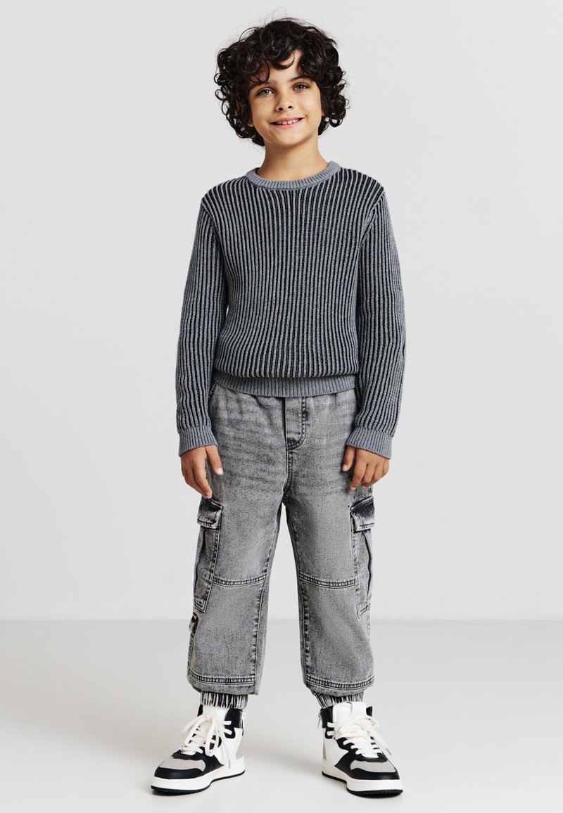 Maglione a strisce grigie con colletto rotondo, abbinato a pantaloni cargo grigi con tasche. Sneakers alte in bianco e nero.