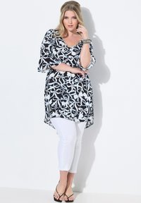 Tunica floreale in bianco e nero con scollatura a girocollo e maniche fluide, abbinata a pantaloni bianchi aderenti e sandali neri con cinturini.