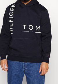 Navy blue hoodie with white text "HILFIGER TOM" on front. Features a kangaroo pocket and drawstring hood. Soft fabric texture.