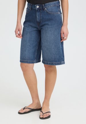 Persoon draagt knielange blauwe denimshorts, zwarte slippers, zwart topje en zilveren ringen aan beide handen, staand tegen een effen achtergrond.