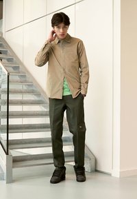 Camisa de botón marrón claro, camiseta verde debajo, pantalones verde oscuro, zapatillas negras. El modelo se encuentra junto a una escalera con barandilla de cristal en un entorno minimalista.