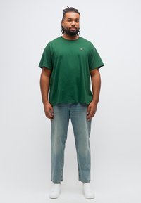 Άνδρας με πλεξούδες, φορώντας πράσινο T-shirt, ανοιχτό μπλε τζιν και λευκά αθλητικά παπούτσια, στέκεται μπροστά σε απλό λευκό φόντο.