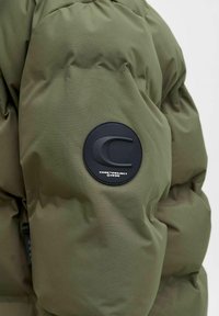 Olivgrön vadderad jacka med quiltade sektioner, en cirkulär svart logotypmärke på ärmen och en dragkedjedetalj vid mudden.