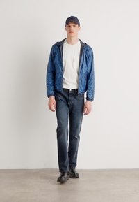 Leichte blaue Jacke über einem weißen T-Shirt, kombiniert mit dunklen Jeans und schwarzen Schuhen; Modell steht vor einem schlichten Hintergrund.