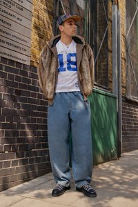 Man in een bontjacket, wit sportshirt met nummer 18, baggy jeans, zwarte schoenen en een geruite pet die op een stoep staat naast een bakstenen muur en een bord.