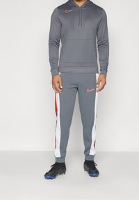 Hombre vestido con un chándal gris de Nike con franjas blancas y rojas en los pantalones y zapatillas de fútbol negras de Nike, de pie frente a un fondo claro y simple.