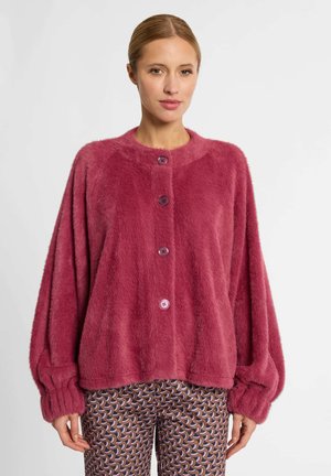 Frau trägt eine lockere, geknöpfte, langärmelige, flauschige rosa Strickjacke und gemusterte braune und rosa Hosen und steht vor einem schlichten Hintergrund.