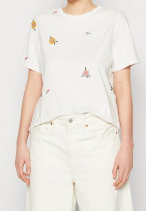 Femme portant une chemise blanche à manches courtes avec de petites broderies florales colorées et un pantalon blanc taille haute, debout devant un fond uni.