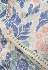 Tissu blanc avec imprimé floral bleu et rose, détails découpés œillets, boutons blancs et bordure brodée décorative.