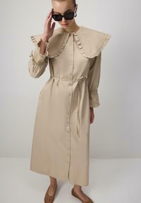 Abito beige con colletto oversize arricciato, maniche lunghe e chiusura a bottoni. Viene fornito con una cintura abbinata e orlo svasato. Indossato con occhiali da sole scuri.