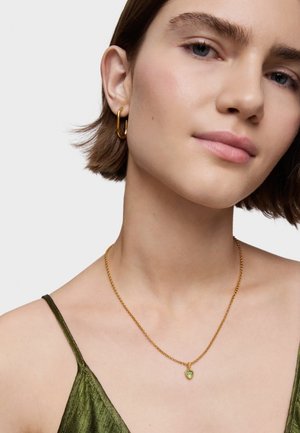 Złoty naszyjnik z sercowym zielonym wisiorkiem. Modelka ma na sobie zieloną satynową bluzkę i złote kolczyki w kształcie kół. Gładka tekstura skóry.