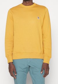 Sudadera amarilla de cuello redondo con un cuello y puños acanalados. Presenta un pequeño logo bordado en el pecho. Tejido suave y texturizado.