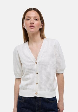 Femme portant un cardigan blanc texturé à manches courtes boutonné avec un col en V festonné et un jean bleu foncé, debout devant un fond uni.