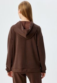 Sudadera con capucha marrón y un acabado texturizado. Diseño holgado, hombros caídos y cinturilla ancha. Simple y funcional.
