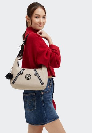 Borsa a spalla beige con due tasche con cerniera, logo rotondo e accenti neri. Indossata con un maglione rosso e una gonna di jeans. Texture liscia.