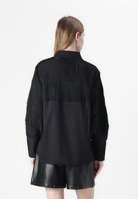 Camisa preta com detalhes de franjas nos ombros e parte superior das costas, combinada com calções de couro pretos brilhantes.