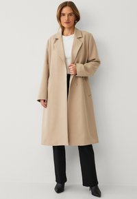 Beige wollen mix jas met notch-kraag, lange mouwen, zijzakken en een enkele knoopsluiting, gecombineerd met zwarte broeken en hakken.