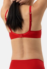 Punainen rintaliivi säädettävillä olkaimilla ja hakaskiristyksellä. Sujuva kangas, jossa on minimalistinen muotoilu, ilman kuvioita tai yksityiskohtia.