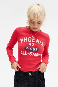 Ungdom med kort blondt hår iført en rød langermet skjorte med teksten "PHOENIX AZ. DIV. 61 ALL-STARS" og mørke dongeribukser.