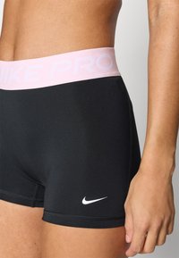 Čierne atletické šortky vyrobené z elastického materiálu s svetloružovým elastickým pásom s textom "NIKE PRO" a bielym logom Nike na nohe.