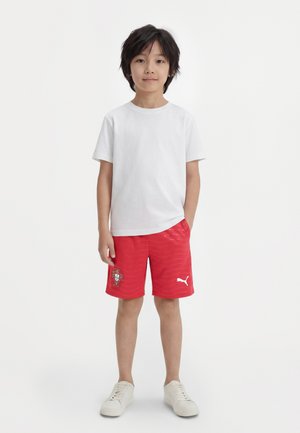 Αγόρι με μαύρα μαλλιά, που φοράει απλό λευκό μπλουζάκι, κόκκινα αθλητικά σορτς με λογότυπα Puma και Πορτογαλίας, και λευκά αθλητικά παπούτσια, στέκεται με τα χέρια στις τσέπες.
