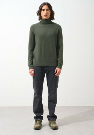 Suéter de cuello alto verde oliva de tela suave, combinado con jeans gris oscuro y zapatillas verdes. Diseño sencillo con una silueta ajustada.