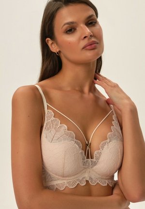 Lace bra in nude, featuring scalloped edges, a deep V-neck design, and a decorative gold accent at the center front. Adjustable straps.