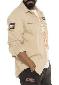 Chemise beige à boutons avec double poche, présentant un patch rayé sur la manche gauche et un T-shirt graphique en dessous. Coupe décontractée et ample.