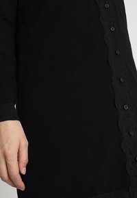 Gros plan sur le torse et la main d'une personne portant un vêtement noir à manches longues avec un devant boutonné orné de dentelle noire.