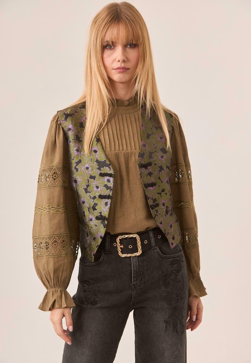 Blusa verde oliva con textura acanalada, superpuesta con un chaleco estampado en verde oscuro y diseño floral púrpura; combinada con jeans negros y un cinturón ancho.