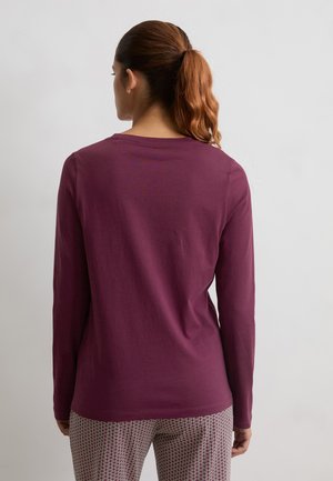 Mujer con pelo castaño en una coleta, vistiendo una camiseta de manga larga morada y pantalones con estampado, de pie contra una pared clara y lisa, de espaldas.