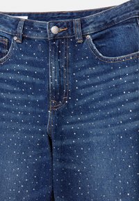 Jean en denim bleu avec de petites clous argentés répartis uniformément sur le tissu et des détails visibles des poches avant et des boutons.